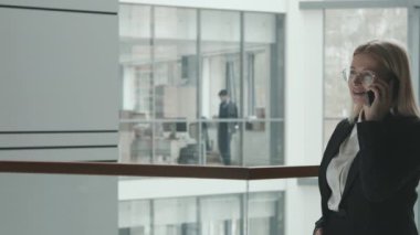 Resmi giyimli ve gözlüklü orta yaşlı sarışın kadın avukatın PAN orta yavaşlığı modern ofiste korkulukların yanında telefon görüşmesi yapıyor.