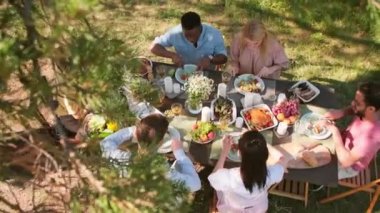 Çok ırklı bir grup gencin parkta ahşap masada oturup leziz bir öğle yemeği yerken çekilmiş en tepeden aşağı görüntüleri.