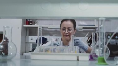 Beyaz önlüklü genç kadın kimyagerin laboratuvar araştırması üzerinde çalışırken mavi çözeltiye baktığı yavaş çekim.