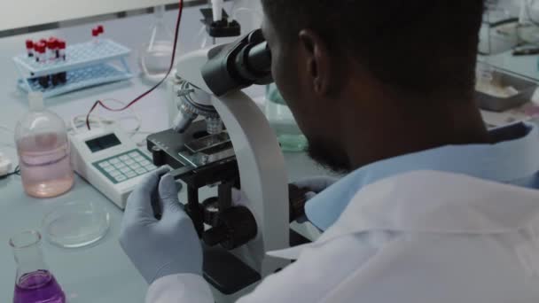 白衣を着た黒人男性科学者の肩のショットと研究室で働いている間 顕微鏡を見て手袋の上に手をスローモーを開催 — ストック動画