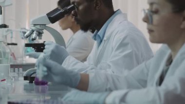 Beyaz önlüklü ve eldivenli Afrika kökenli Amerikalı erkek bilim adamlarının yavaş bakış açısıyla mikroskop kullanarak, sonra kameraya doğru dönüp, yanında oturan ve araştırma yapan kadın meslektaşlarına doğru dönün.