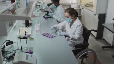 Beyaz önlüklü, eldivenli ve yüz maskeli bilim adamlarından oluşan yavaş bir ekibi yukarı kaldır. Laboratuvarda araştırma yapıyorlar. Engelli kadın kimyagerler tekerlekli sandalyede çözümü deniyorlar.