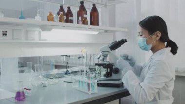 Yüz maskeli, eldivenli ve beyaz önlüklü Asyalı kadın bilim adamının yavaş çekimleri raflarda mavi solüsyonla dolu test tüpünü yerleştiriyor ve laboratuvarda çalışırken mikroskop kullanıyor.