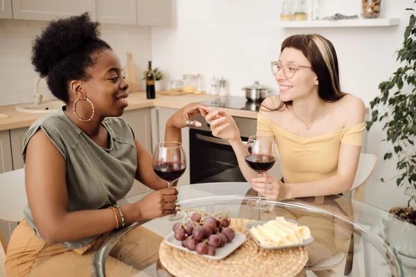 Deux petites amies heureuses avec des verres de vin rouge assis dans la cuisine — Photo