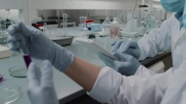 Beyaz önlüklü ve eldivenli, tablet üzerinde notlar alan ve laboratuvar araştırmaları yaparken test tüpünde çözeltiyi inceleyen tanınmamış bilim insanlarının el kamerasıyla yakından çekilmiş fotoğrafı.