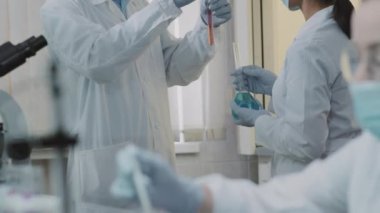 Beyaz önlüklü, eldivenli ve yüz maskeli Afrikalı Amerikalı erkek bilim adamı ve Asyalı bayan meslektaşının yavaş yavaş öne eğilmeleri ve şişe ve test tüplerinde çözümleri tartışmaları.