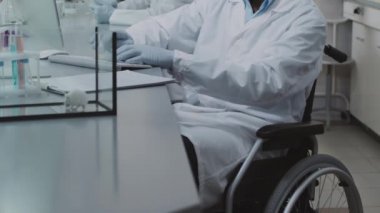 Beyaz önlüklü, eldivenli, engelli erkek bilim adamlarının laboratuvarda bilgisayar üzerinde çalışırken laboratuar çalışanlarının laboratuvar farelerini camdan bir tankta beklemelerini istiyorum.