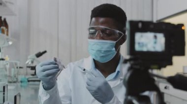 Yüz maskeli, eldivenli ve beyaz önlüklü siyah bilim adamlarının yavaşlaması beyaz laboratuvar faresi tutuyor ve öğrenciler için video çekerken ve talimatlar verirken ona deneysel ilaç enjekte ediyor.