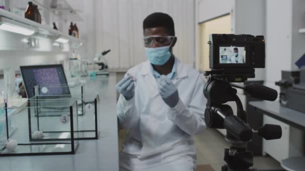 ビデオカメラを録画して話をする前に座っている白いコート フェイスマスクで黒人男性科学者の遅い中出し その後実験薬でラットを注入 — ストック動画