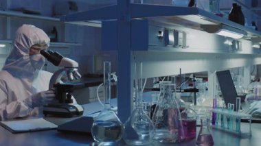 Tek kullanımlık tulumlu ve yüz maskeli bilim adamlarının yavaş PAN 'ı mikroskop altında örnekler üzerinde çalışıyor ve laboratuvarda dizüstü bilgisayarda yazı yazıyor.