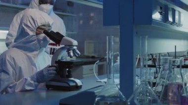 Koruyucu tulumlar, gözlükler, yüz maskesi ve eldivenlerle laboratuvar meslektaşı erkek arkadaşında mikroskop kullanan ve içi kan dolu test tüpleriyle konuşan tanımlanamayan kadın bilim adamının yavaş yavaş PAN 'ı.