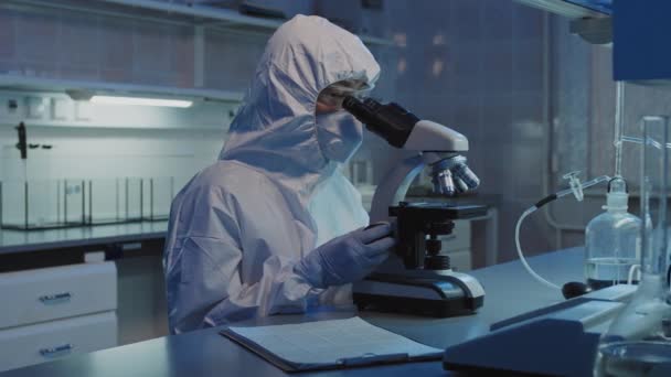 研究室の顕微鏡下でサンプルを見て保護カバー ゴーグル フェイスマスクで認識できない疫学者のスローモパンショット — ストック動画