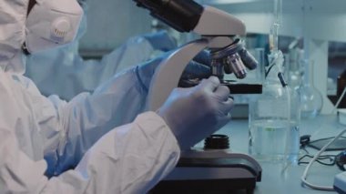 Tek kullanımlık tulumlu, yüz maskeli, gözlüklü ve eldivenli tanımlanamayan siyah erkek epidemiyoloji uzmanının, laboratuarda mikroskop altında cam kaydırmasıyla yavaş çekim yapın.