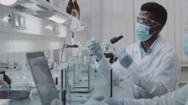 Beyaz önlüklü, gözlüklü ve yüz maskeli Afrikalı erkek bilim adamlarının PAN yavaş yavaş beyaz laboratuvar faresine deneysel ilaç enjekte ederken Asyalı bayan meslektaşı dizüstü bilgisayar üzerinde çalışıyor.