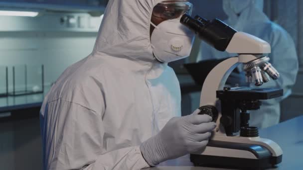 在实验室显微镜下 将无法辨认的身穿工作服 面罩和护目镜的非洲裔美国男性科学家的慢镜头倾斜 在显微镜下观察样品 然后看着相机 — 图库视频影像
