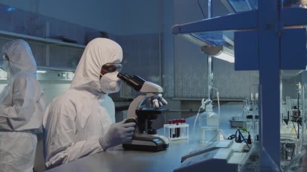 Pan拍摄的无法辨认的身穿工作服 护目镜 手套和面罩的非裔美国男性科学家坐在实验室柜台前 使用显微镜 然后看着相机 — 图库视频影像
