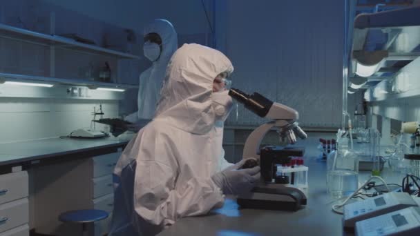 使い捨てカバーでアジアの女性疫学者のPanの低速 研究室で顕微鏡を使用してゴーグルや顔マスク その後 カメラを見て — ストック動画