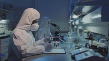 Tek kullanımlık tulumlu, gözlüklü ve yüz maskeli iki epidemiyolojistin laboratuvar tezgahında oturup kan örneklerini incelerken yavaş çekim görüntüleri.