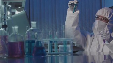 Koruyucu tulumlu, gözlüklü, eldivenli ve yüz maskeli kadın bilim adamlarının yavaş yavaş test tüplerini mor solüsyonla incelemesi