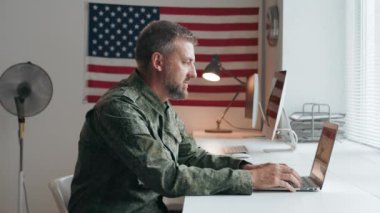 Orta yaşlı, askeri üniformalı bir Amerikan subayının portresi masada oturuyor ve dizüstü bilgisayarda yazıyor, sonra kameraya doğru dönüyor ve poz veriyor.