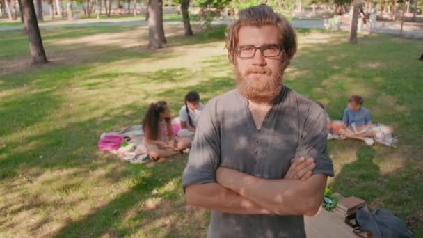 晴れた夏の日に公園で腕を組んで立っている眼鏡のひげを生やした男性教師の肖像画を追跡し カメラを見て 背景に毛布の上に座っている小学生のグループ — ストック動画