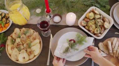 Kadın ellerinin, peçete, çam dalı ve böğürtlenlerle süslenmiş tabakla Noel masasını hazırlayışının en üst görüntüsü. Biberiyeli kızarmış patates, cevizli fırında armut, ekmek ve meyve suyu masada.
