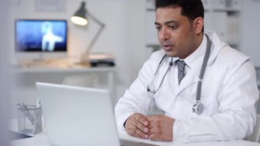 Orta Doğulu pratisyen hekimin laptopun önünde oturup hastayla görüntülü konuşma yaparken çekilmiş yatay portresi.