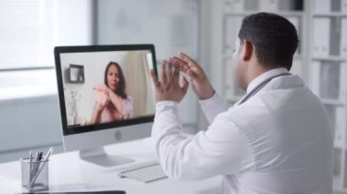 Profesyonel pratisyen hekim ofisinde oturmuş video başında kadın hastayla çalışıyor.