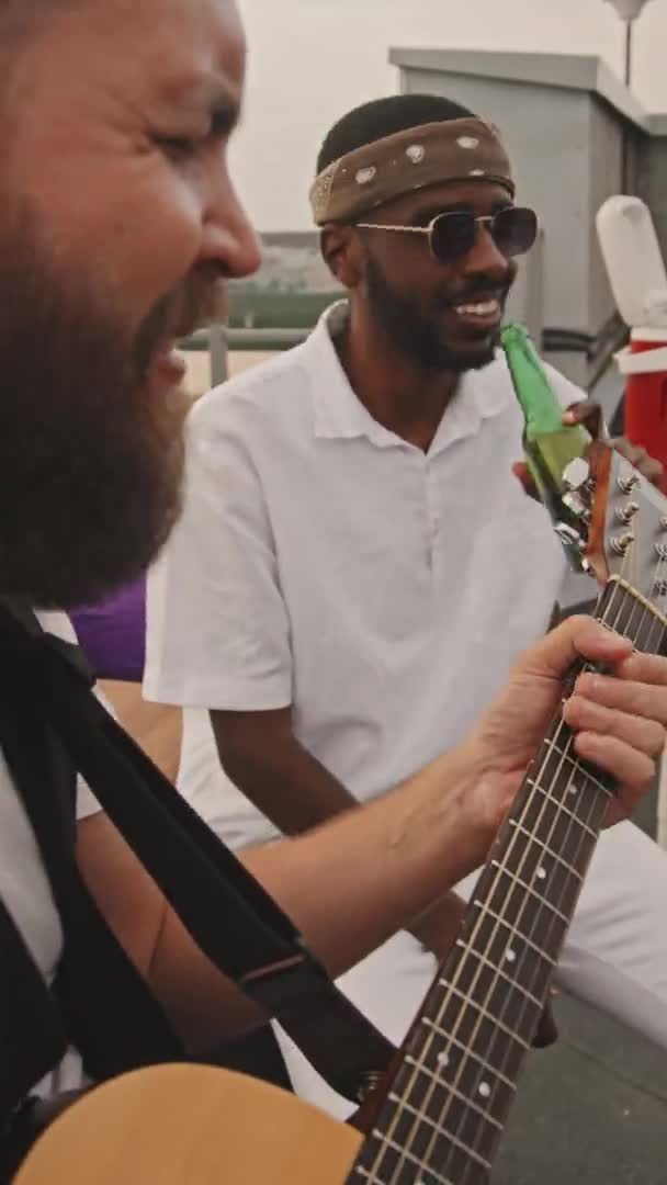 陽気な髭を生やした白人青年がアコースティックギターを弾きながら笑顔で アフリカ系アメリカ人男性の友人がビールを飲みながら屋上パーティーで音楽を楽しんでいます — ストック動画