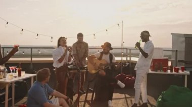 Çatıdaki terasta, rüzgârlı bir yaz akşamında müzik çalan bandonun el kameralı görüntüsü.