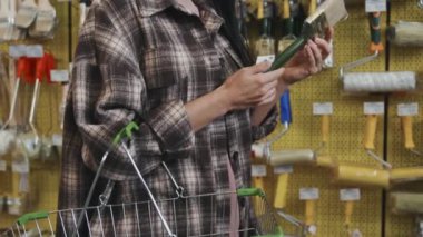 Nalburdaki rafların önünde duran ve boya fırçasına bakan güzel ve genç bir kadının yan görüntüsüyle yukarı doğru eğil.