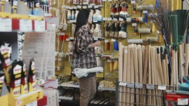 Elinde alışveriş sepeti olan genç bir kadının hırdavatçıdaki rafların önünden geçerken el kamerasıyla fırçayı eline alıp kalitesini kontrol etmesi.