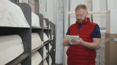 Sakallı, kabarık yelek giyen, tablet kullanan ve ev yapımı mağazadaki duvar kâğıdı rulolarını kontrol eden yavaş erkek amir.