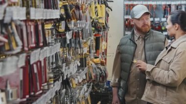 Hırdavatçıdaki rafların önünde duran ve malzemeleri seçerken konuşan kadın ve erkeğin yavaş çekimleri.