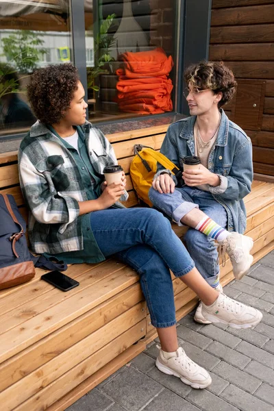 Подростковая пара в повседневной одежде сидит на скамейке у современного здания — стоковое фото