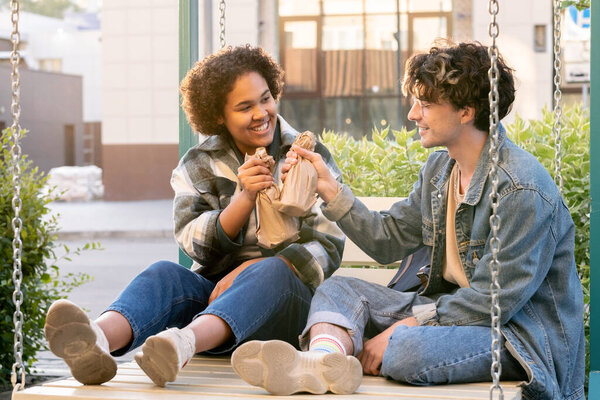 Casual teens with bottles wrapped into paper relaxing on swings