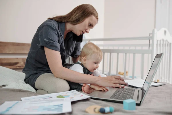 Poważna Młoda Matka Siedzi Łóżku Papierami Pomocą Laptopa Podczas Gdy — Zdjęcie stockowe