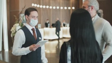 Genç erkek otel müdürünün yavaş çekimleri. Yüz maskeli. Misafirleri odalarına götürmeden önce dijital tablet kullanarak lobiye giriş yapıyorlar.