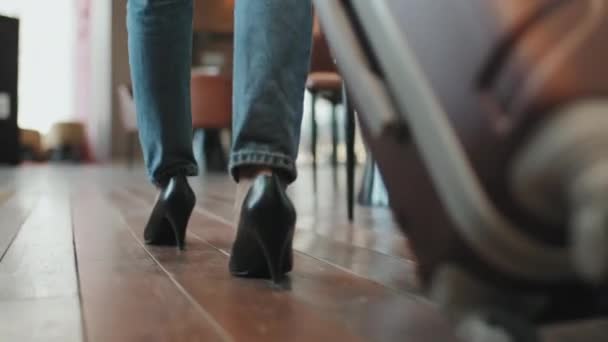 低セクションのバックビュースローモの認識できない女性のジーンズやヒールを着てホテルのロビーを歩くスーツケースを運ぶ — ストック動画