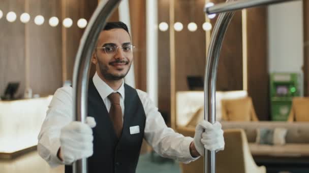 中低速肖像画の笑顔若い男性ベルボーイで制服姿でカメラのために荷物カート立っているでPoshホテルのロビー — ストック動画