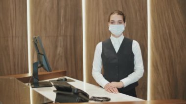 İş yerinde kameraya poz veren üniformalı ve maske takan kadın otel resepsiyonistinin orta yavaş portresi.