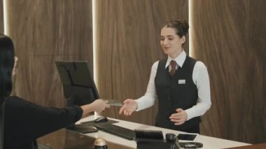 Genç bayan resepsiyonistin, yeni müşteriyi karşılarken, pasaportunu alırken ve modern birinci sınıf otellerde boş oda ararken orta boy fotoğrafı.