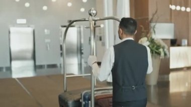 Otel lobisinden asansörlere bavul taşıyan üniformalı bir erkek hamalın dikiz görüntüsü.
