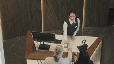 Modern çiftlerin oteldeki yavaş çekimleri sırasında masanın yanında duran genç bayan resepsiyonistle konuşarak odalarına pasaport ve otel kartı vererek iyi tatiller dileklerini iletiyorlar.