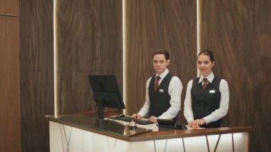 Modern yüksek sınıf lobide kameraya poz veren iki genç erkek ve kadın otel yöneticisinin ya da üniformalı resepsiyonistin orta yavaş portresi.