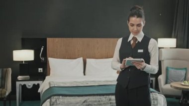 Modern konforlu otel odasında elinde dijital tabletle kameraya gülümseyen üniformalı kadın otel müdürünün orta yavaş portresi.