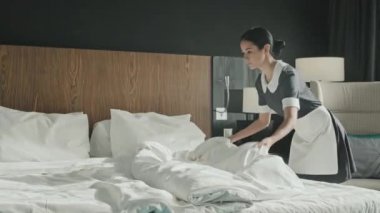 Orta boy, modern lüks otel odasında beyaz çarşaf değiştiren üniformalı melez hizmetçi.