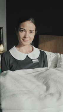 Modern otel odasında dururken elinde taze beyaz havlularla kameraya gülümseyen genç melez hizmetçinin dikey portresi.