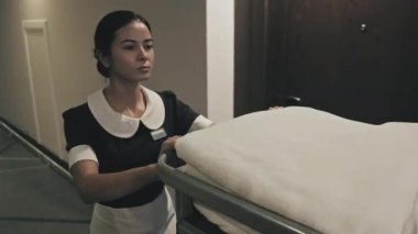 Otel koridorunda yürürken kadın hizmetçinin havluları, çarşafları ve temizlik malzemelerini el arabasında taşırken görüntüsü.