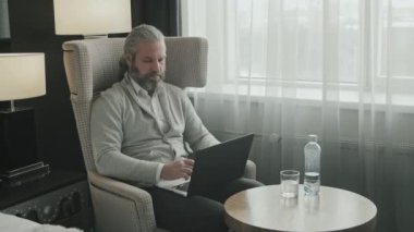 Modern otel odasındaki koltukta oturmuş dizüstü bilgisayarla çalışan başarılı bir iş adamının orta boy fotoğrafı.